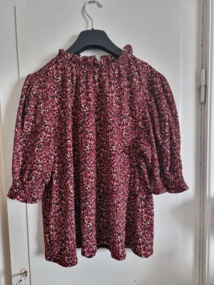 Blommig blus från Holly & Whyte st M  - Söt blommig blus från Holly & Whyte med puffiga trekvartsärmar och volang vid halsen. Färgen är vinröd med små vita, rosa och svarta blommor. Tillverkad i ett lätt och luftigt tyg, perfekt för en trendig och avslappnad look.