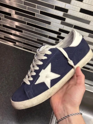 Golden goose skor - Snygga blå sneakers från Golden Goose med vit stjärndetalj på sidan och vita skosnören. Skorna har en låg profil och vit sula med vintage-look. Ovandelen är i mocka och detaljer i läder. Perfekta för dig som gillar streetstyle och unika detaljer.