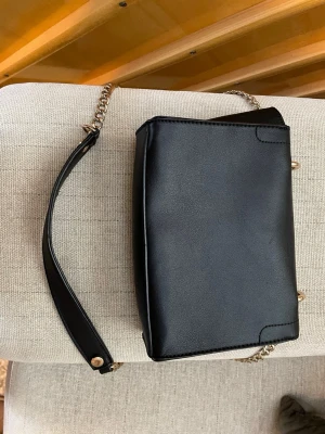 Svart axelremsväska med kedja - Snygg svart handväska i slätt konstläder med guldfärgad kedjerem och detaljer. 19 cm bred. 6 cm djup, ca 15 cm hög. Aldrig använd. 