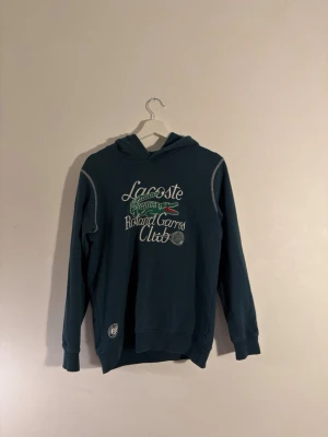 Grön Lacoste hoodie Roland Garros - Mörkgrönt hoodie från Lacoste med Roland Garros-tryck och den klassiska krokodilloggan framtill. Hoodien har huva, vita kontrastsömmar vid axlarna och muddar vid ärmslut och nederkant. 