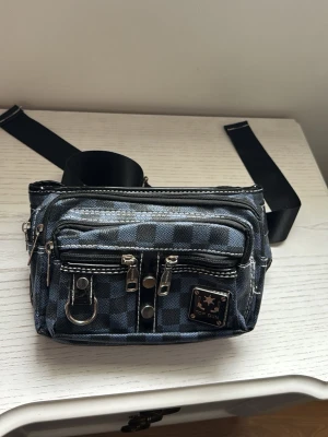 Rutigt midjeväsk från Top Gun - Snygg midjeväska från Top Gun i svart och grått rutigt mönster. Väskan har flera dragkedjefack, silverfärgade detaljer och justerbart band med spänne. Perfekt för att bära det viktigaste och hålla händerna fria.