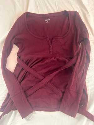Vinröd ribbad topp från Hollister - Supersnygg vinröd långärmad topp från Hollister med ribbad struktur och knappar framtill. Toppen har omlottdetalj med knytband i midjan och en rundad halsringning. Perfekt för dig som vill ha en trendig och bekväm look. (Pris kan diskuteras 💕)