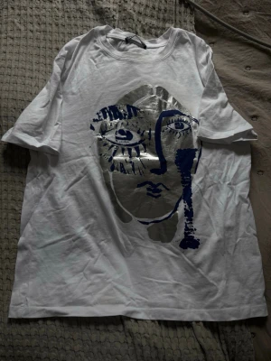 Vit t-shirt med konstnärligt tryck - Vit t-shirt med ett stort konstnärligt ansiktstryck i silver och mörkblått på framsidan.