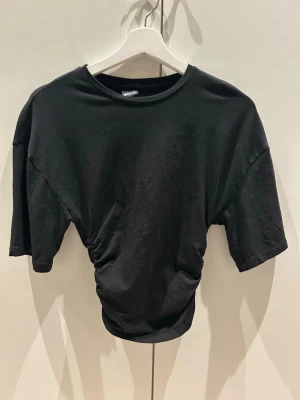 Svart topp  - Trendig svart croppad topp med korta ärmar och snygg rynkdetalj i midjan. Perfekt till höga jeans eller kjol. Mjuk och skön i bomullsmaterial med avslappnad passform och rund halsringning.