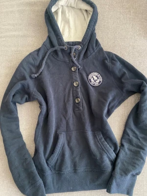 Mörkblå hoodie från Abercrombie & Fitch - Mörkblå hoodie med knappar framtill och dragsko i huvan från Abercrombie & Fitch. Broderad logga på bröstet och känguruficka. Insidan av huvan är ljus och mjuk. Perfekt för en avslappnad och cool stil. Det står storlek M på tröjan men jag tycker att den passar xs/s. Måtten: längd 55 cm bredd 48 cm