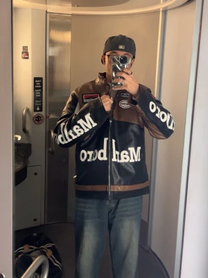 Svart och brun Marlboro skinnjacka - Säljer en svart och brun skinnjacka från Marlboro med stora vita logotexter på bröstet och ärmarna. Jackan har dragkedja framtill, två fickor med dragkedja och flera patchar, bland annat från Michelin och Alpinestars. Snygg retro racingstil.