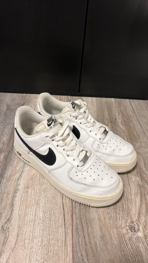 Nike Air Force 1 vita sneakers - Klassiska vita Nike Air Force 1 sneakers med svart swoosh på sidan. Skorna har perforerad tå, vita skosnören och en robust sula med AIR-detalj. Tillverkade i läder med textildetaljer och låg profil som ger en clean och tidlös look.