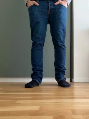 Nudie Grim Tim Jeans 32/31 - Säljer ett par klassiska blå jeans från Nudie Jeans med smal passform och fem fickor. Jeansen har orangea kontrastsömmar, normal midja och är tillverkade i denim. Perfekta för dig som gillar en enkel och stilren look. Skonsamt använda och inga defekter.