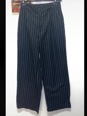 Svarta kostymbyxor med vita ränder - Säljer ett par svarta kostymbyxor med vita smala ränder. Byxorna har vida/raka ben, hög midja och hällor för skärp. Kan bäras som lågmidjade byxor beroende på din storlek. 