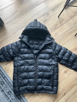  Peak Performance Svart dunjacka  - Snygg svart dunjacka från Peak Performance med huva och diskret logga på bröstet och ärmen. Jackan har quiltad design, dragkedja framtill och elastiska muddar vid ärmslut och midja. Perfekt för kalla dagar och riktigt skön att bära.