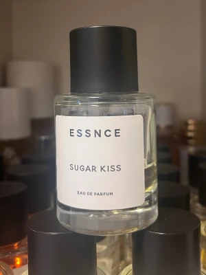 ESSNCE Sugar Kiss Eau de Parfum - ESSNCE Sugar Kiss Eau de Parfum kommer i en stilren, rund glasflaska med svart lock och vit etikett. Doften har en modern och minimalistisk design som passar perfekt på sminkbordet. Flaskan är genomskinlig och etiketten har enkel svart text.