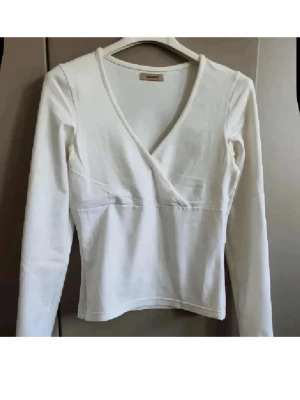 Vit omlottopp från Vero Moda - Säljer en stilren vit omlottopp från Vero Moda. Toppen har lång ärm, v-ringad omlottdesign och är tillverkad i mjuk bomull. Perfekt att matcha med jeans eller kjol för en clean look.