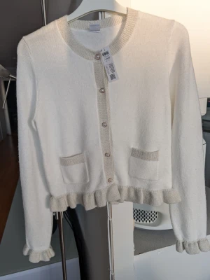 NY Vit kofta med volangkant och fickor - Supersöt vit kofta med beige detaljer, volangkant nertill och vid ärmslut. Storlek 146/152 från Lindex. Två små fickor framtill och rund halsringning. Knäpps med pärlemorknappar. Oanvänd, lappar kvar.