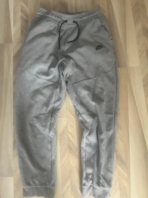Nike tech byxor - Snygga grå mjukisbyxor från Nike med svart logga på vänster sida. Byxorna har dragsko i midjan och muddar vid bensluten för en skön passform