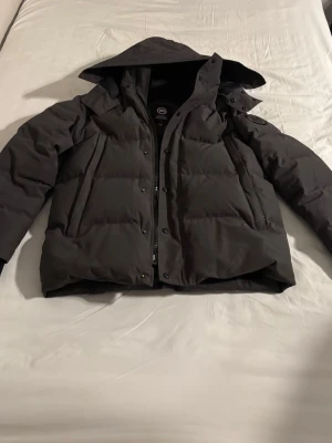 Canada goose black label - Snygg svart dunjacka från Canada Goose med huva och dragkedja framtill. Jackan har en quiltad design och praktiska fickor på sidorna. Perfekt för kalla vinterdagar och håller dig varm med sin tjocka vaddering. Ett litet hål vid armhålan men det går att sy.