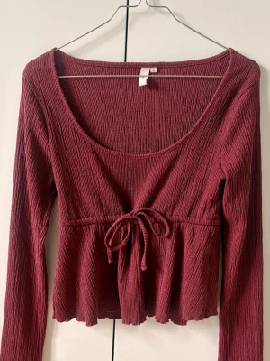 Vinröd croppad topp med knyt - Trendig vinröd croppad topp med lång ärm och snygg knytning framtill. Toppen har en ribbad struktur och lätt volangkant nedtill ♥️