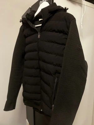 Svart jacka från Moncler - Snygg svart pufferjacka från Moncler med quiltad front och huva. Ärmarna är ribbade och har en diskret Moncler-logga på vänster ärm. Jackan har dragkedja framtill och två sidofickor med dragkedja. Avtagbar luva också. Använt den kanske 4 gånger enbart