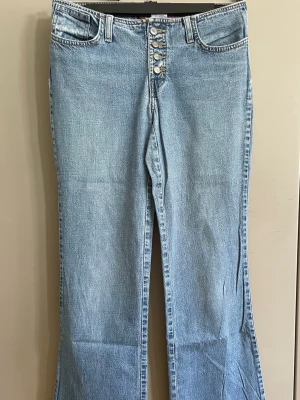 Ljusblå bootcut jeans med knappar - Säljer ett par ljusblå bootcut jeans med hög midja och fem knappar framtill. Klassisk femficksmodell i mjuk denim med lätt utsvängda ben. Perfekta för dig som gillar retrovibbar och avslappnad stil.midjemått 79/80 cm 