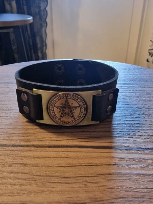 Armband vintage pentagram - Svart läderarmband med ett detaljerat pentagram i metall. Armbandet har justerbara tryckknappar för att passa olika handledsstorlekar. Storlek 23 cm långt. Bredd 3 cm.