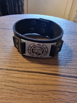 Armband vintage horus öga - Svart läderarmband med ett detaljerat hörus öga i metall. Armbandet har justerbara tryckknappar för att passa olika handledsstorlekar. Storlek 23 cm långt. Bredd 3 cm.