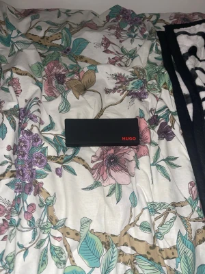 Svart glasögonfodral från Hugo - Snyggt svart glasögonfodral från Hugo med röd insida och logga i hörnet. Fodralet är hårt och skyddar dina glasögon på ett stilrent sätt. Perfekt accessoar för dig som vill ha koll på dina brillor och samtidigt hålla det classy.