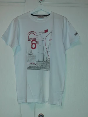 Vit t-shirt Regatta med segelbåtar - Säljer en vit t-shirt från Regatta med tryck av segelbåtar och texten 'Antigua 6th Edition' i rött och grått på framsidan. T-shirten har rund hals och korta ärmar. Perfekt för dig som gillar marina vibbar och stilrena tryck. (Storlek S för man, M för kvinna.)