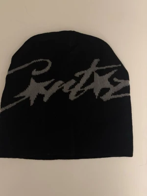 Svart mössa med grått tryck - Cool svart mössa med stort grått logotyptryck i grafisk stil över hela framsidan. Mössan är slätstickad och har en enkel, rund form som passar perfekt till streetwear och vardagsstil. 