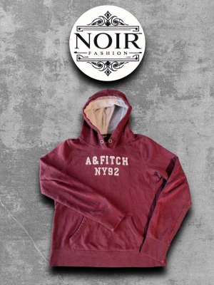 Vinröd hoodie från A&Fitch - Snygg vinröd hoodie från A&Fitch med vit text 'A&FITCH NY92' på bröstet. Hoodien har en stor huva med ljus insida och klassiska kängurufickor framtill. Perfekt för en avslappnad och trendig stil.