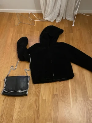 Svart tjejfluffjacka från Only S med en stilig handväska tiamo (Bundle) - Mysig svart teddyjacka med huva och dragkedja framtill. Jackan har en fluffig och mjuk struktur som ger en cool och avslappnad look. Perfekt för kyliga dagar när du vill vara både varm och trendig.