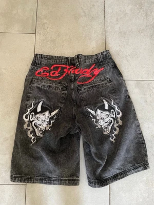 Svarta Ed Hardy shorts med demonbrodyr - Unika svarta jeansshorts från Ed Hardy med grå tvätt och stora broderade demonmotiv på bakfickorna. Röd Ed Hardy-logga både fram och bak. Klassisk femficksmodell med loose fit och hög midja. Perfekta för dig som vill sticka ut.
