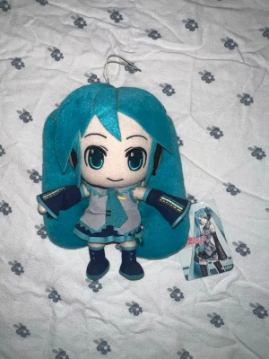 BNWT Hatsune Miku japansk plushie - Säljer en helt ny med lappen kvar mjuk plush av Hatsune Miku från Crypton Future Media importerad från Japan. Dockan har turkos hår i två långa tofsar, detaljerad outfit med grå topp, blå kjol och svarta skor. Perfekt som accessoar till maskerad eller cosplay. Materialet är mjukt tyg och fyllning. Ungefär 20cm lång. 