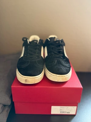 Svarta sneakers från Valentino - Snygga svarta sneakers från Valentino med vit sula och vit sidopanel. Skorna har en klassisk låg profil, svarta skosnören och en unik kamouflagemönstrad yttersula. Tillverkade i mocka och läder för en exklusiv känsla.