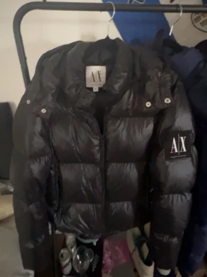 Svart pufferjacka från Armani Exchange - Snygg svart pufferjacka från Armani Exchange med stor logga på ärmen. Jackan har hög krage, dragkedja framtill och quiltad design som ger en riktigt trendig look. Perfekt för kalla dagar när du vill hålla stilen.