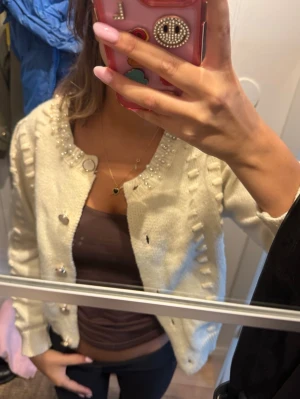 Vit kofta med pärldetaljer - Söt vit kofta med glittriga pärlor och dekorativa detaljer längs knäppningen och halsringningen. Koftan har silvriga knappar och ribbade muddar vid ärmslut och nederkant. Perfekt för dig som vill ha en extra touch till din outfit.