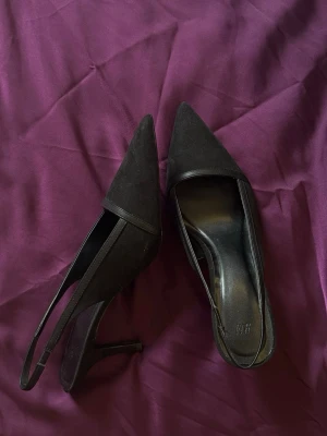 Svarta slingback pumps från H&M - Stilrena svarta pumps från H&M med spetsig tå och smal klack. Skorna har öppen häl med slingback-rem och är tillverkade i mockaimitation. Perfekta för dig som gillar en klassisk och elegant look.