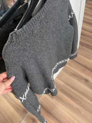 Grå stickad tröja från ONLY - Mysig grå stickad tröja från ONLY med vita korsstygnsdetaljer runt ärmar och nederkant. Tröjan har rund halsringning och långa ärmar, perfekt för att hålla sig varm under kyliga dagar. Materialet är mjukt och ger en avslappnad look.