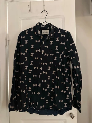 Svart skjorta med mönster på flugor från Our Legacy i storlek 46/S - Snygg svart skjorta från Our Legacy med vitt abstrakt mönster över hela plagget. Skjortan har klassisk krage, knäppning framtill och långa ärmar.   100% bomull. Sparsamt använd, i fint skick!