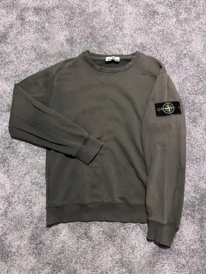 Olivgrön sweatshirt från Stone Island - Säljer en olivgrön sweatshirt från Stone Island med den klassiska logotypen på vänster ärm. Tröjan har rund halsringning, ribbade muddar och är tillverkad i mjuk bomull. Perfekt för en avslappnad och stilren look. Enda nackdelen är att tvättlappen är borta!