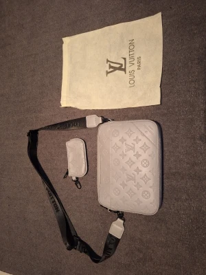 Grå axelväska från Louis Vuitton - Snygg grå axelväska från Louis Vuitton med präglat monogrammönster och justerbar svart axelrem. Väskan har en rektangulär form och kommer med en matchande liten accessoarväska. Tillverkad i slätt läder och levereras med dustbag.