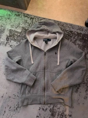 Grå hoodie från Polo Ralph Lauren - Tjocka snören, röd logga och grå zip. Riktigt fin, behöver tvättas då dene lite smutsig vid handen