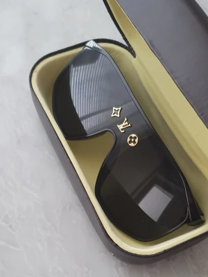 Svarta solglasögon - Säljer ett par svarta solglasögon från Louis Vuitton med breda bågar och guldfärgade LV-detaljer framtill. Glasögonen har en modern, kantig form och kommer i originalfodral. Perfekta för dig som vill ha en lyxig och trendig accessoar.