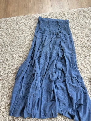 Blå långkjol från Ezence - Säljer en blå långkjol från Quinte med smockad midja och volangdetaljer. Kjolen har en luftig och avslappnad passform, perfekt för dig som gillar en bohemisk stil. Materialet känns mjukt och lätt, vilket gör den skön att bära under varma dagar.