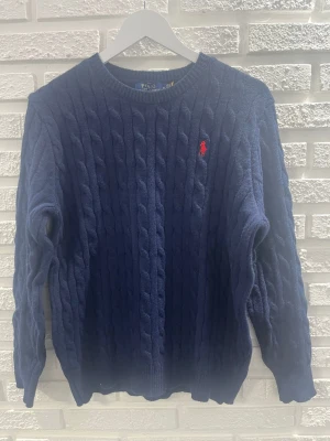 Mörkblå kabelstickad tröja Polo Ralph Lauren - Mörkblå kabelstickad tröja från Polo Ralph Lauren med klassisk rund halsringning och röd broderad logotyp på bröstet. Tröjan har långa ärmar och ribbade muddar. Perfekt för att hålla sig varm och snygg under kyliga dagar.