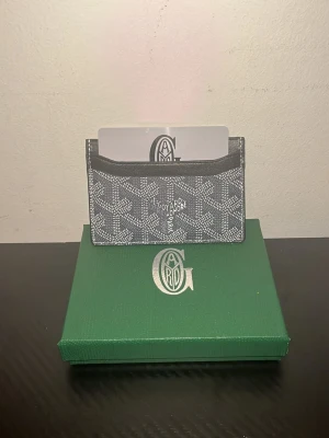 Goy4ard grå kortfodral korthållare - Snyggt grått kortfodral från Goy4ard med det klassiska mönstret i vitt. Tillverkad i canvas med läderdetaljer och har flera fack för kort. Kommer med originalask och dustbag. Perfekt för dig som vill ha en stilren och exklusiv accessoar.