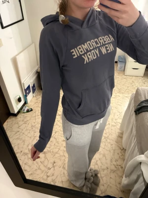 Blå hoodie Abercrombie New York - Mysig blå hoodie från Abercrombie & Fitch med trycket 'Abercrombie New York' i vitt på bröstet. Hoodien har huva och en stor magficka framtill. Perfekt för chill dagar och enkel att matcha med andra plagg.