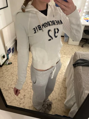 Vit hoodie från Abercrombie & Fitch - Vit hoodie med blå text och siffran 92 från Abercrombie & Fitch. Klassisk modell med huva och dragsnören, ribbade muddar och magficka. Perfekt för en avslappnad och sportig stil. Mjuk och skön bomullsblandning.