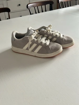 Gråa Adidas sneakers med vita detaljer - Snygga gråa Adidas sneakers med klassiska vita ränder på sidorna och vita skosnören. Skorna har en låg profil och är tillverkade i mocka med gummisula. Perfekt för dig som gillar en clean och sportig look.