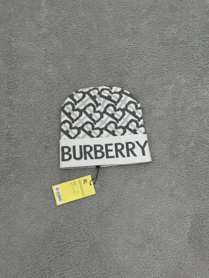 Vit och grå Burberry mössa - Säljer en snygg vit och grå mössa från Burberry med ikoniskt monogrammönster och stor logga framtill. Mössan är ribbstickad och har en uppvikt kant. Perfekt accessoar för dig som vill ha en trendig och exklusiv look.