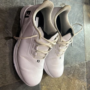 Vita golfskor från FootJoy - Säljer ett par vita golfskor från FootJoy med klassisk snörning och diskret logga på tungan och hälen. Skorna har en sportig design med slitstark ovandel och vadderad insida för extra komfort. Perfekta för dig som vill ha stilrena och funktionella skor på golfbanan.