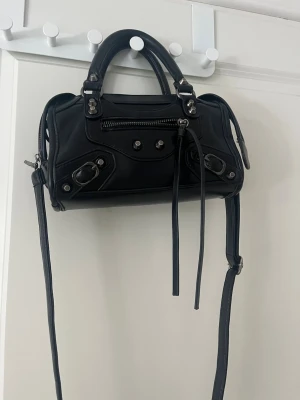 Balenciagaliknande svart handväska i läder - Säljer en svart handväska lik Balenciagaväskan i mjukt läder med silverfärgade metalldetaljer. Väskan har både handtag och en lång axelrem, samt en dragkedjeficka framtill och dekorativa spännen. Perfekt för dig som gillar en edgy och trendig look.